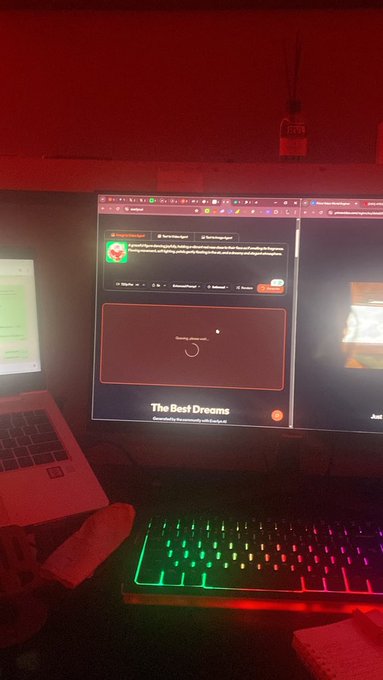 A desk setup illuminated in red light featuring a monitor showing a music player interface with 'The Best Dreams' displayed, an RGB keyboard, and a laptop to the side.