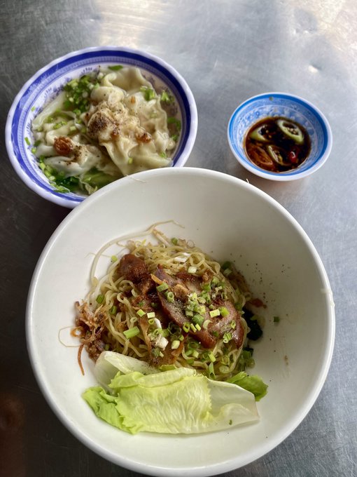 A meal of noodles topped with meat and lettuce in a white bowl, served with wontons in broth and a small dish of soy sauce on a metal table.
