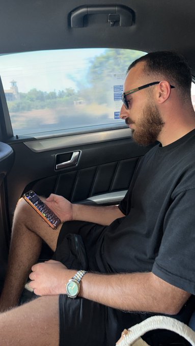 A bearded man wearing sunglasses sits in the back of a moving car, focused on his phone which displays a chat interface, with a luxury watch on his wrist and the blurred scenery of a coastal road passing by outside the window.