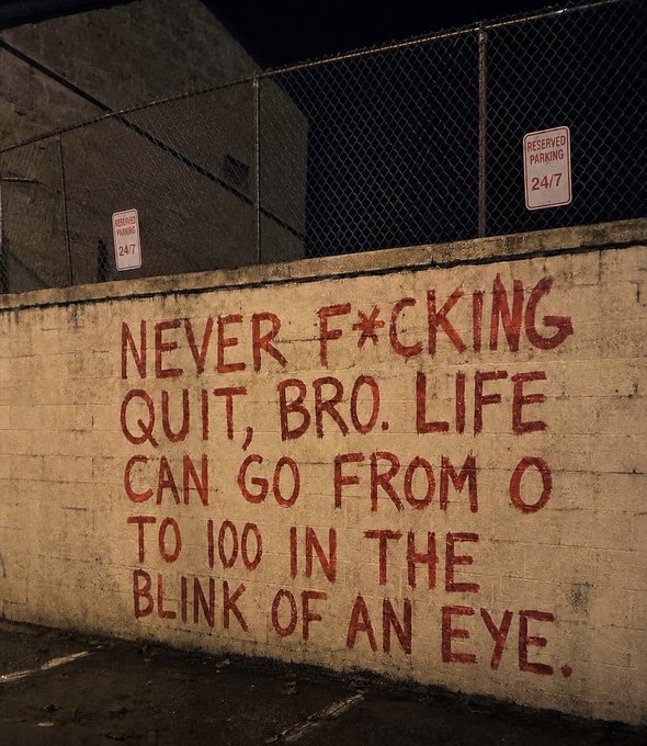 A nighttime photograph of a concrete wall with red graffiti reading 'NEVER F*CKING QUIT, BRO. LIFE CAN GO FROM 0 TO 100 IN THE BLINK OF AN EYE,' behind a chain-link fence with 'RESERVED PARKING 24/7' signs.