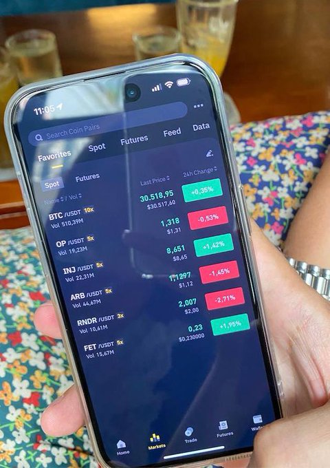 A person holding a smartphone displaying a crypto trading app showing BTC at $30,518.95, along with OP, INJ, ARB, RNDR, and FET prices with 24h changes.