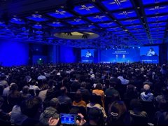Wide shot of a large indoor convention hall filled with attendees seated facing a stage with blue lighting and screens, likely an anime or pop culture event.