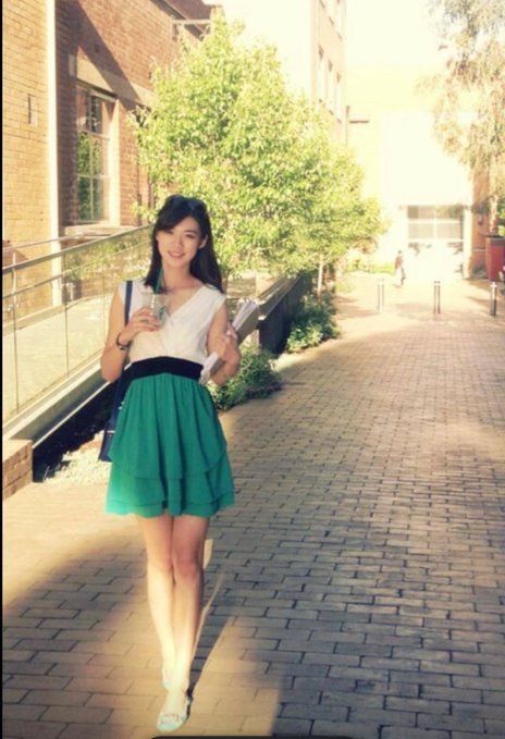 A young woman wearing a white top and a green ruffled skirt, walking on a brick pathway in what appears to be a campus or urban setting with greenery.