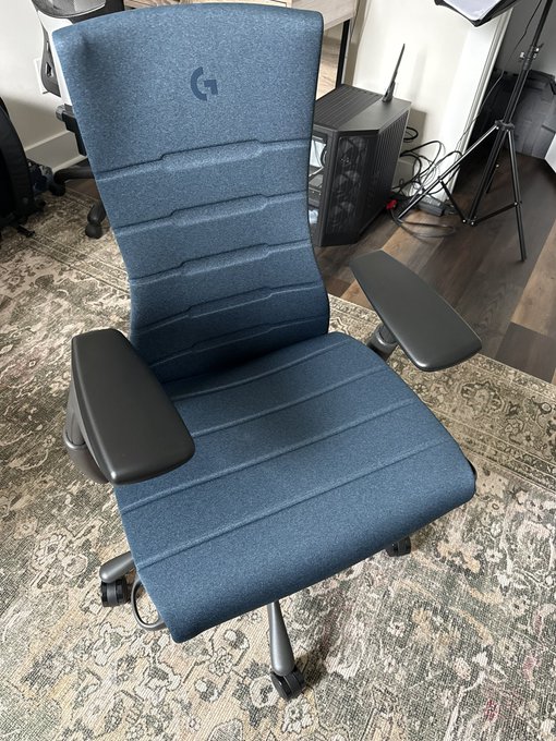 A blue gaming chair with the Logitech G logo on the backrest sits on a patterned rug in a room with a gaming PC and tripod in the background.