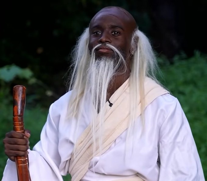 A portrait of an older Black man with a long white beard and white robes, holding a wooden staff while standing outdoors in a natural setting.