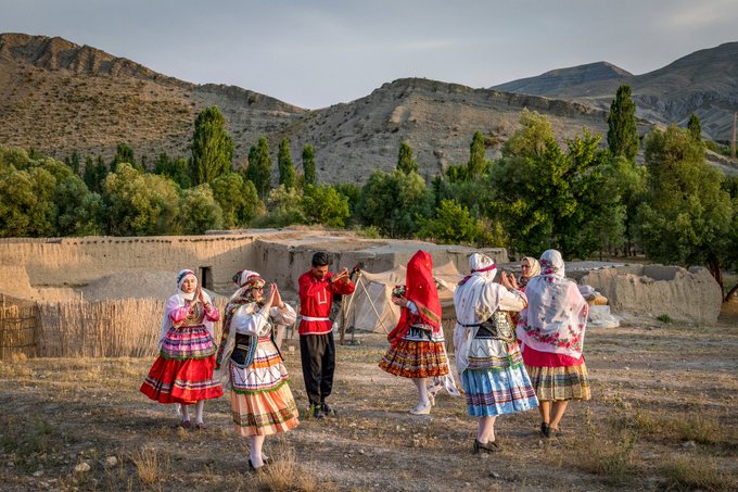 A group of people in traditional folk costumes dancing in a rural village with mud-brick houses, set against a backdrop of arid hills and green trees.