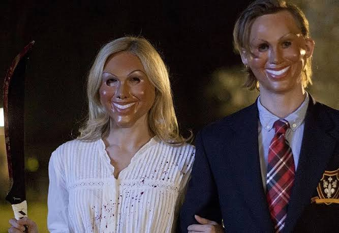 Two people wearing unsettling smiling face masks, one in a white blouse and one in a navy suit with a red plaid tie and crest, pose together at night.