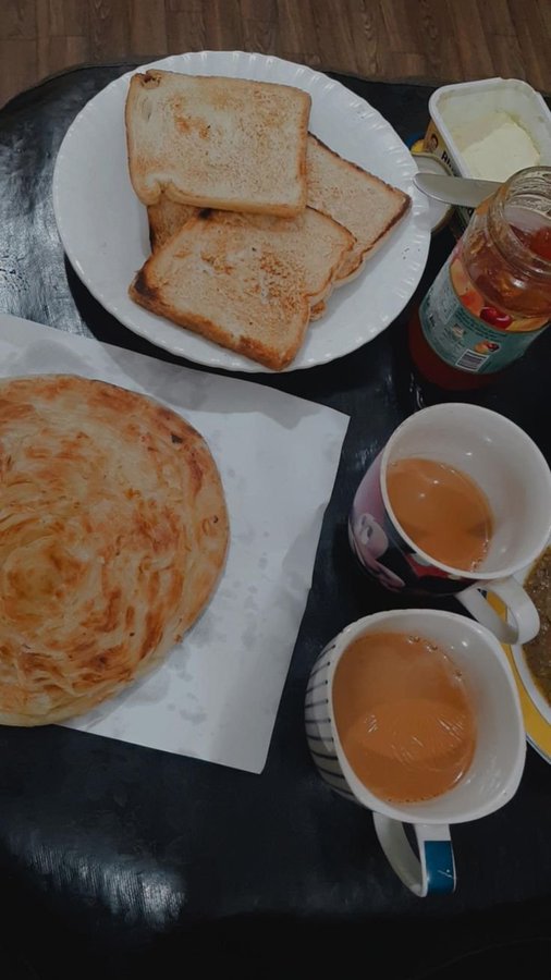 A breakfast spread on a dark table includes toasted bread on a white plate, a layered pastry, two mugs of milky tea, and a jar of jam or spread.
