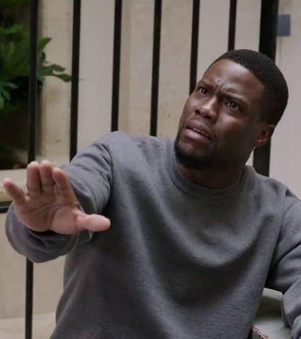 A close-up image of Kevin Hart looking exasperated or defensive, with his hand raised in a 'stop' gesture, wearing a gray sweatshirt.