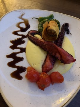 A plated gourmet dish featuring what appears to be octopus tentacles over a pool of yellow sauce, garnished with cherry tomatoes, a rolled vegetable, and drizzled with a dark balsamic reduction.
