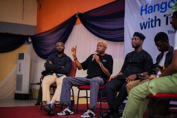 A photograph of a panel discussion with four people seated on chairs on a stage with draped fabric, where one man in the center is speaking into a microphone.