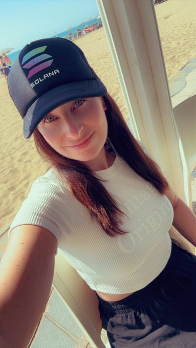 A selfie of a young woman wearing a navy 'SOLANA' baseball cap and a white 'Authentic Origin' t-shirt, posing on a sandy beach under a clear sky.