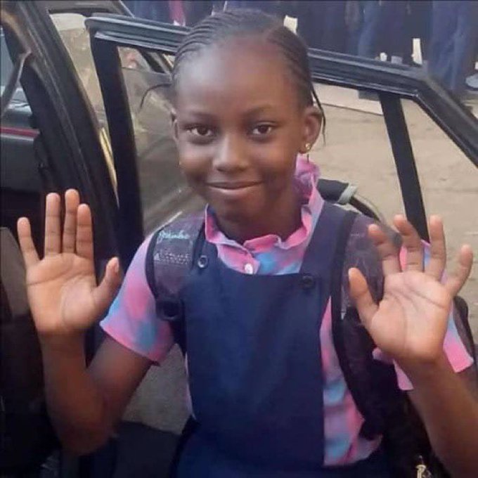 A young girl in a school uniform with a pink and blue plaid shirt smiles and holds up both hands with palms facing the camera.