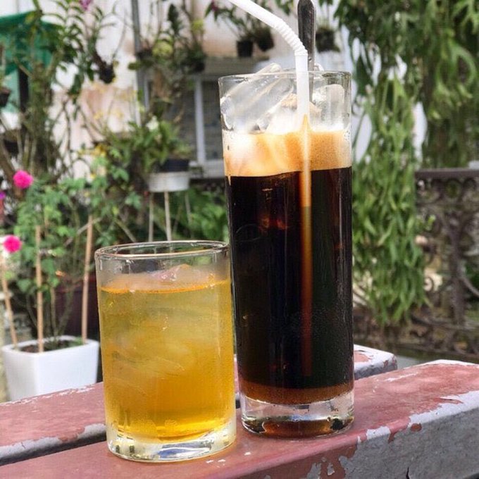 Two iced beverages on a red railing: a short glass with a yellow drink and a tall glass with a dark soda containing a straw, with a garden of green plants and pink flowers in the background.