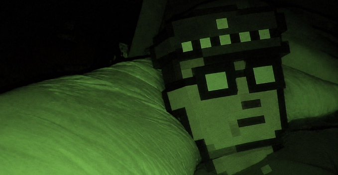 A pixelated plush toy resembling a Minecraft-style character rests on a pillow in a dimly lit room bathed in green light, capturing the cozy, spooky aesthetic of "Basetober."