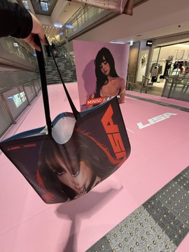 A black handbag featuring a Lisa portrait print is displayed in a pink-themed retail showcase, with a matching Lisa poster in the background.
