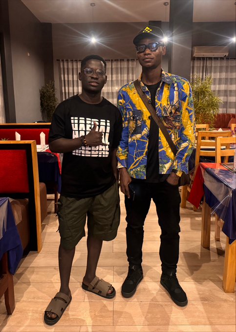 Two young men posing indoors in what appears to be a restaurant; the left one wears a black graphic t-shirt and green shorts, while the right one wears glasses, a cap, a yellow and blue patterned jacket, and black pants.