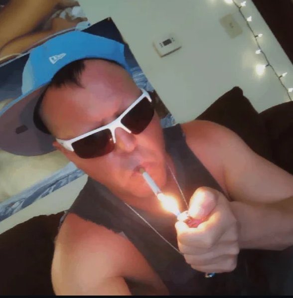 A man wearing a backwards blue cap and white sunglasses lights a cigarette in a dimly lit room.