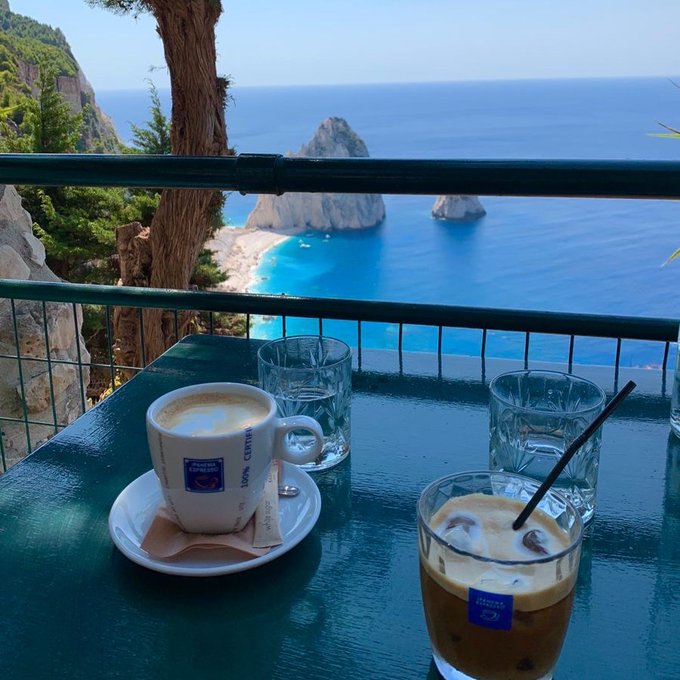 A table setting with a cup of espresso and a glass of iced coffee on a green table, overlooking a stunning turquoise ocean and rocky coastal scenery.