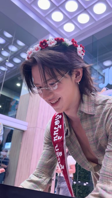 A smiling person with glasses wearing a plaid jacket, a flower crown headband, and a red sash with white text.