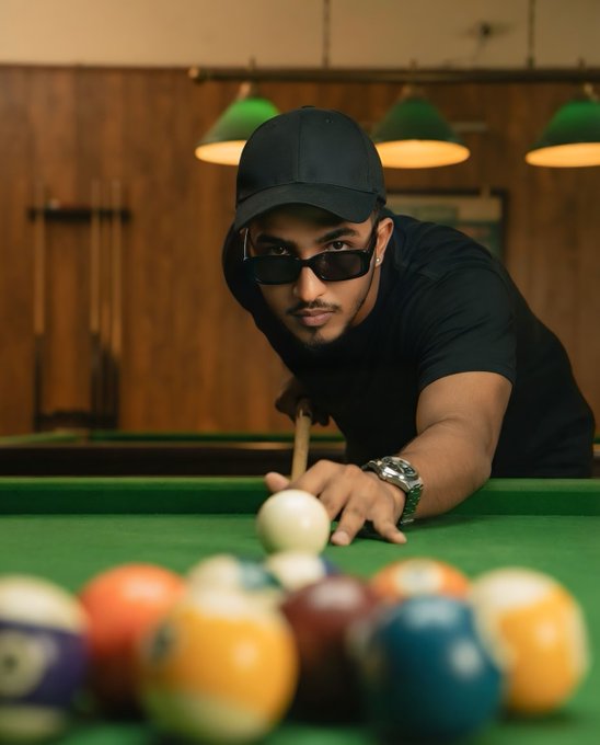 A man in a black cap, dark sunglasses, and black shirt is leaning over a pool table, lining up a shot with a cue stick amid scattered billiard balls.