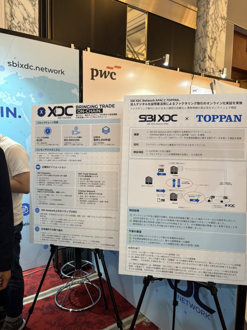 A photograph of a conference display booth featuring 'sbixdc.network' branding and a 'pwc' logo, showcasing presentation boards about 'BRINGING TRADE EDUCATION' and an 'SBIXDC x TOPPAN' pilot project, with Japanese text and diagrams explaining the cross-border trade financing initiative.