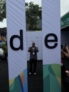 A man wearing a dark polo shirt and lanyard stands with arms raised between two tall banners with the large black letters "d" and "e", at what appears to be an outdoor event.