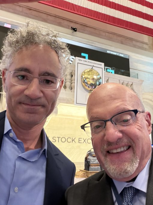 Two men pose for a selfie in front of the New York Stock Exchange podium and bell, with Palantir (PLTR) signage visible in the background, celebrating a visit to the trading floor amid excitement over the stock hitting new highs.