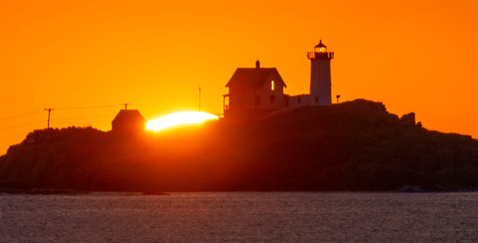 A scenic sunset photograph captures the silhouette of a lighthouse and adjacent buildings on a rocky coastline, with bright orange and yellow light reflecting across the water.