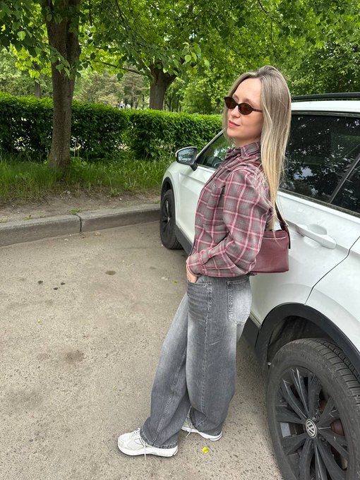 A woman with blonde hair wearing sunglasses, a plaid shirt, and baggy jeans leans against a white car while holding a maroon handbag.