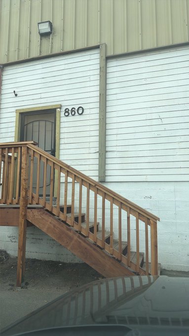 A wooden staircase leading up to a door marked '860' on the side of a white building with green corrugated metal trim.
