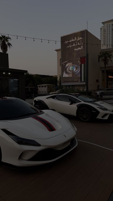 A dusk photo of two white sports cars parked in front of a building displaying a large billboard for 'Celebrity Watch Boutique' in Arabic.
