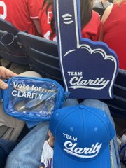 The image features a person sitting in a chair, holding up two blue foam fingers with the word "team clarity" written on them. The person also has a blue baseball cap with the same text on it. In addition to these items, there is a small bag or purse placed next to the person's feet.