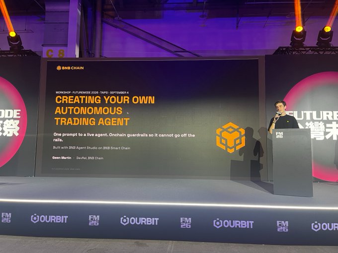 A man stands at a podium presenting a workshop titled 'CREATING YOUR OWN AUTONOMOUS TRADING AGENT' at a BNB Chain event, with 'OURBIT' and 'FM26' logos on the stage.