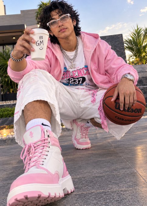 A young man squatting outdoors wearing a pink hoodie, white shorts, and pink sneakers, holding a Wilson basketball and a 'GM' branded cup.