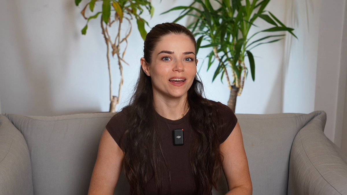 A woman sits on a light gray couch wearing a brown top with a "dji" branded microphone clipped to it, appearing as a guest on the Payward Pulse video podcast. The interview setting with houseplants in the background corresponds to the episode's discussion about Kraken's US stocks launch in the EEA, AnthropicAI's Project Glasswing, and White House/Wyoming appearances by Payward's leadership.