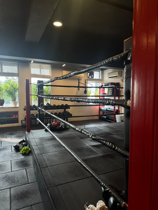 An indoor boxing gym features a boxing ring with black ropes and red posts, with training equipment like gloves and shoes scattered on the floor.