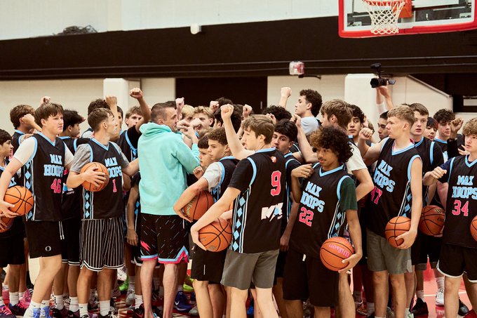 A youth basketball team wearing 'MAX HOOPS' jerseys gathers in a huddle, holding basketballs and raising their fists in a team cheer inside a gymnasium.