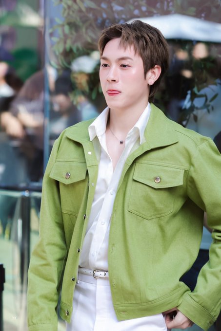A young man wearing a green jacket over a white shirt and white pants, standing outdoors with a blurred background.