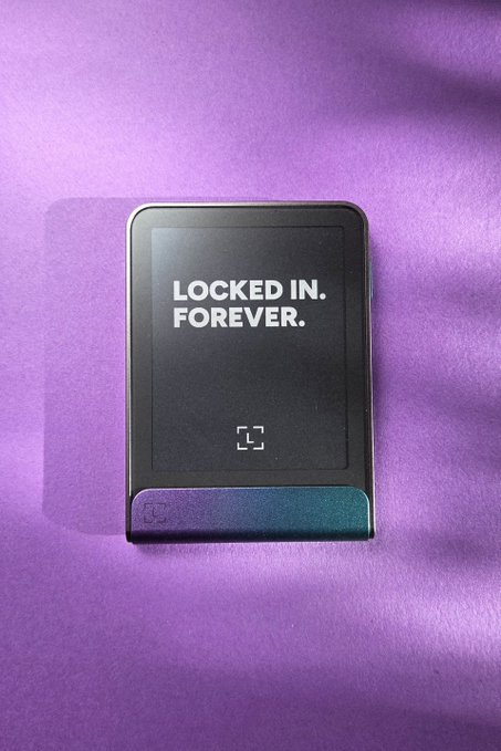 A small black electronic device displaying the text 'LOCKED IN. FOREVER.' on its screen, placed on a purple surface.