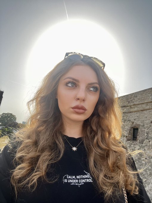 A selfie of a woman with long wavy brown hair wearing sunglasses on her head and a black t-shirt that reads 'CALM, NOTHING IS UNDER CONTROL', photographed with the sun creating a halo effect behind her.