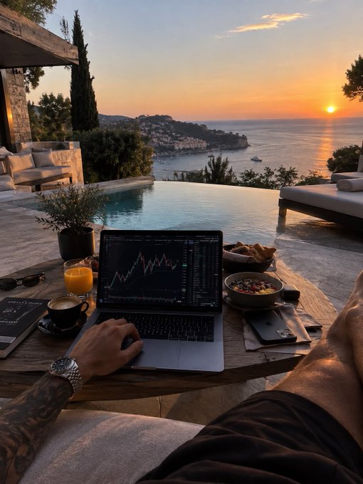 A first-person view of a person with tattoos using a laptop displaying a trading chart, sitting by a pool overlooking the ocean at sunset with breakfast and coffee nearby.