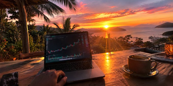 A serene scene shows a person working on a laptop displaying stock market charts on a wooden table, with a cup of coffee nearby, overlooking a beautiful sunset over the ocean.