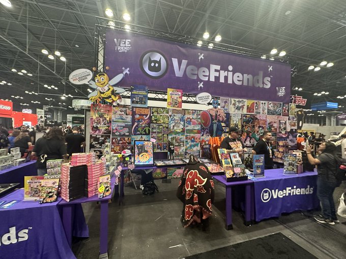 A bustling convention booth displays a wide array of colorful VeeFriends merchandise, including trading cards and figures, with people browsing under the large purple 'VeeFriends' banner.