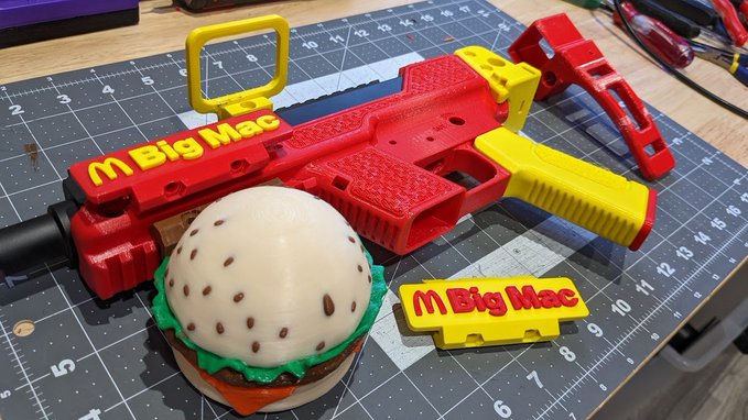 A 3D-printed red and yellow toy gun labeled 'Big Mac' displayed on a cutting mat alongside a printed hamburger prop and a matching 'Big Mac' nameplate.
