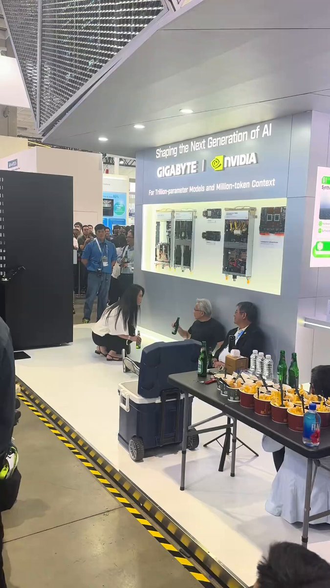 The image shows a group of people gathered around a table at an event, with some sitting down while others stand nearby. The table has several items on it, including bottles and a laptop. A person wearing a suit can be seen standing near the center of the scene. In front of them is a sign that reads "AVGO - 13% AH - NVDA CEO Jensen Huang:".