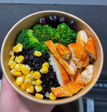 A close-up photograph of a hand holding a paper bowl filled with a meal containing broccoli, dark olives, sweet potato slices, corn kernels, and black rice.