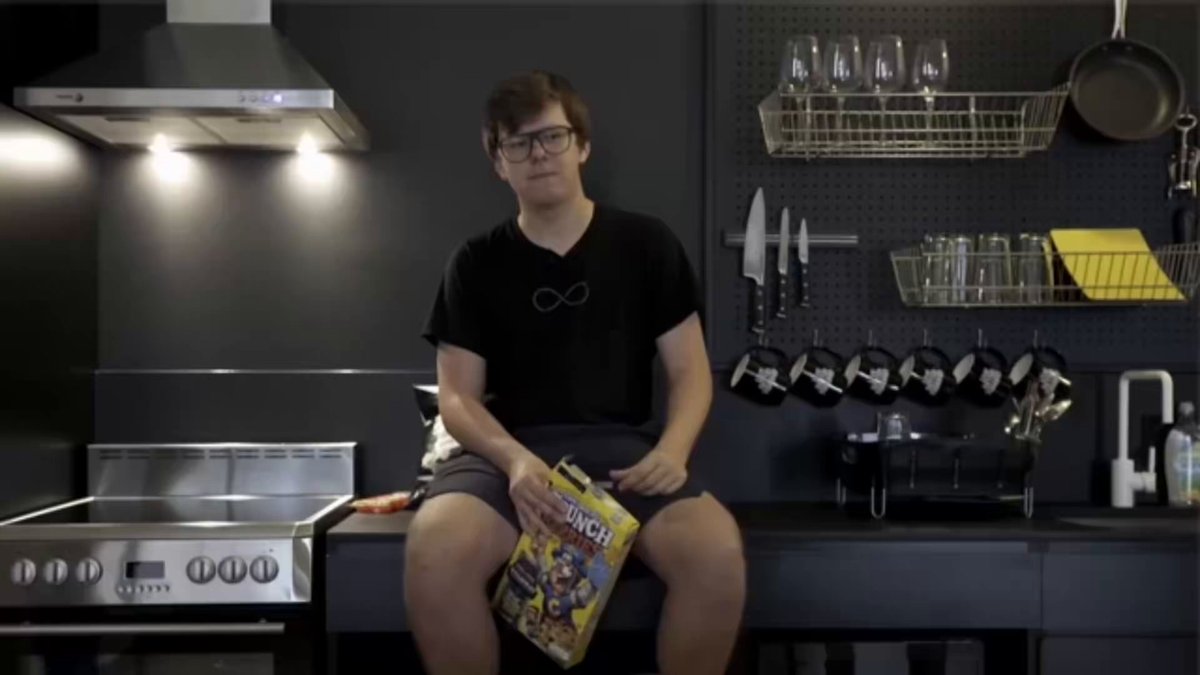 The image features a young man sitting at an oven, holding a magazine or newspaper while wearing glasses. The kitchen has various items such as cups, wine glasses, knives, spoons, and bowls placed around him. There are also two sinks visible in the scene. The background of the image is black with white text that reads "12 YAŞINDAKİ ÇOCUK 1.000 DOLARI BİTCOİN'E YATIRDI, YILLAR SONRA MİLYONER OLDU Hikayemizin kahramanı Erik Finman adında gezinirken keşfettiği bitcoin aldı Yıllar geçti ve bu yatırımı onu milyoner yaptı". The young man is sitting in front of an oven, and the kitchen appears to be well-equipped with various utensils and items.