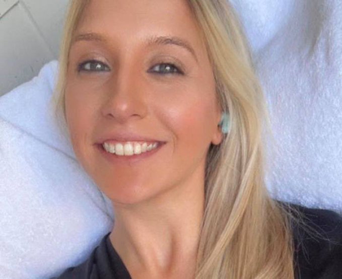 A close-up selfie of a blonde woman smiling, lying against a white pillow or towel.