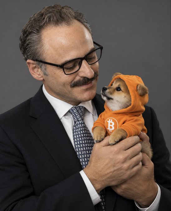 A man with a mustache and glasses in a suit holding a small Shiba Inu puppy dressed in an orange hoodie with a Bitcoin logo.